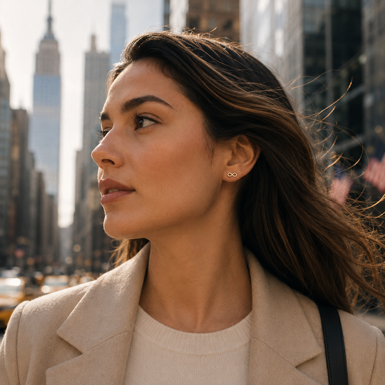 A woman in a beige blazer and cream sweater wearing a small gold infinity-shaped stud earring, photographed outdoors in a city setting with blurred skyscrapers in the background.