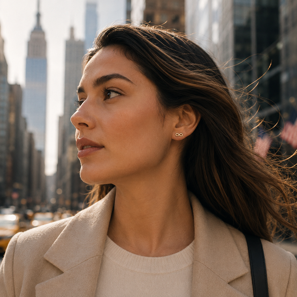 A woman in a beige blazer and cream sweater wearing a small gold infinity-shaped stud earring, photographed outdoors in a city setting with blurred skyscrapers in the background.